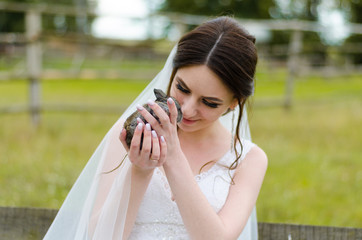 Young woman bride smiling and holding cute rabbit over park summer nature outdoor. White wedding dress, green background