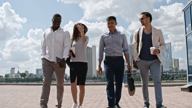 Diverse business team of young colleagues walking outdoors along modern glass building towards the camera, drinking coffee, smiling and chatting