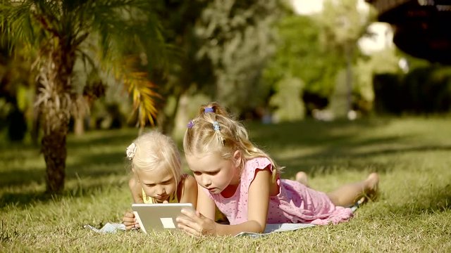 two cute girls play with a tablet computer lying on the grass in the bushes near the palm trees