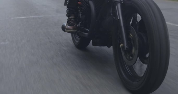 A guy in a black leather jacket and helmet riding a classic motorcycle on a forest road. Close-up from the wheels