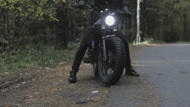 A guy in a black leather jacket and helmet riding a classic motorcycle on a forest road.
