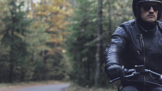 A guy in a black leather jacket and helmet riding a classic motorcycle on a forest road.