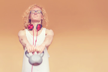Skinny curly blond man in a white vest and with red headphone lifting dumbbell on a yellow background