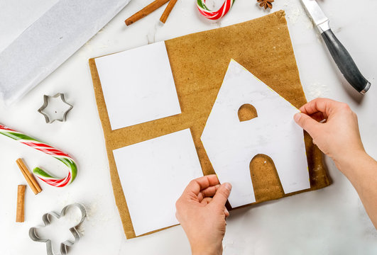 Preparing for Christmas, a person, hands in the frame, makes a gingerbread house. Cutting on a paper stencil from the dough, baking tray, decoration in the frame. On a white table, top view copy space