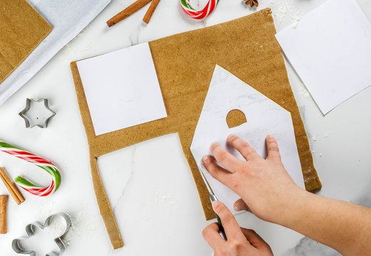 Preparing for Christmas, a person, hands in the frame, makes a gingerbread house. Cutting on a paper stencil from the dough, baking tray, decoration in the frame. On a white table, top view copy space