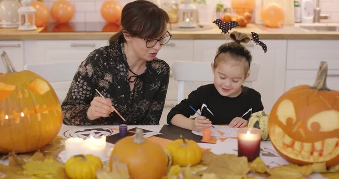 Mother and daughter in cute Halloween costume sitting at table near pumpkins and leaves and making Halloween decorations.