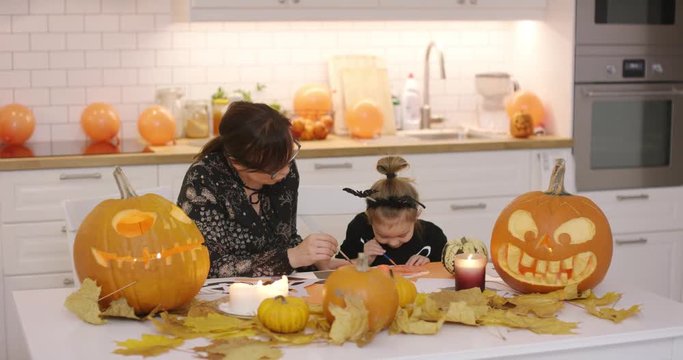 Woman and her cute little daughter in Halloween costume sitting at table near jack-o-lanterns and leaves and painting Halloween decorations.
