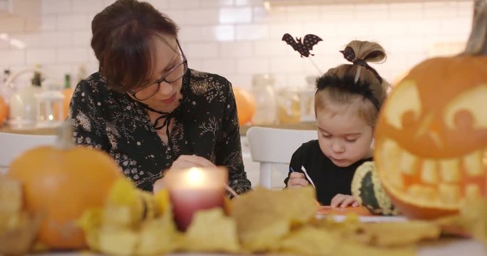 Woman in glasses and her daughter in Halloween costume sitting at table and making decorations for Halloween party.