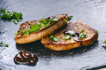 Grilled pork chops, steaks with vegetables, coriander, beans and sauce on a black slate. Fresh meat with foam. Dark background. Herbs