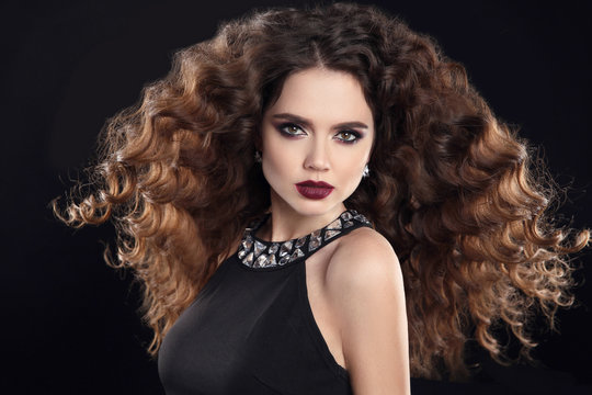 Hairstyle. Fashion Brunette Girl With Long Curly Hair, Beauty Makeup. Glamour Portrait Of Beautiful Woman With Marsala Matte Lips, Blowing Hairstyle Isolated On Black Background.