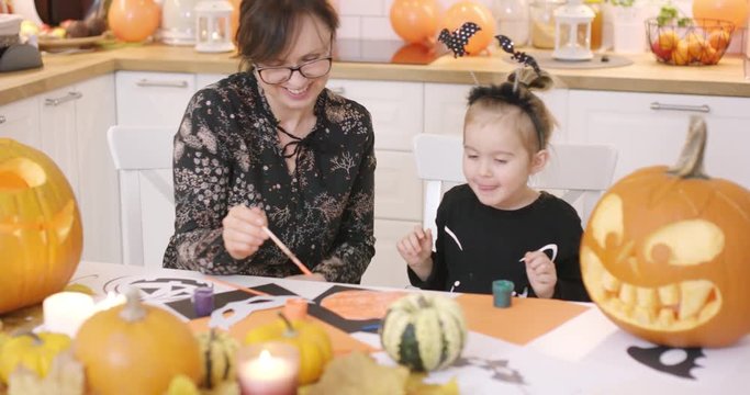 Mother and her cute little daughter sitting at kitchen table near jack-o-lantern and other Halloween decorations and looking at painted pumpkin.