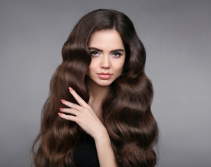 Beauty hair. Brunette girl with long shiny wavy hair. Beautiful model portrait with curly hairstyle and eyeliner makeup isolated on studio gray background.