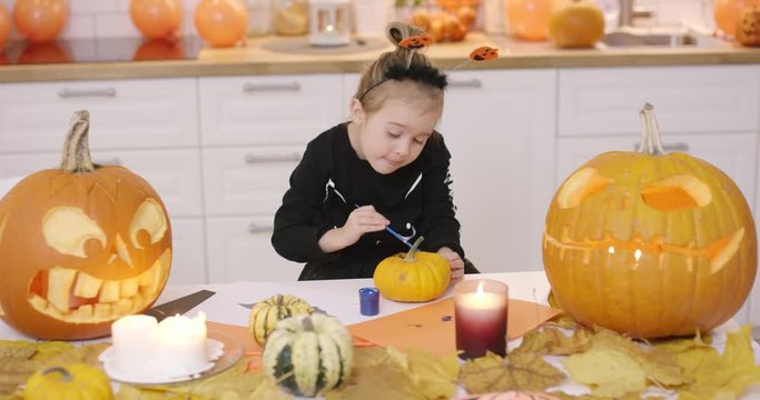 Little girl in cute Halloween costume standing near kitchen table and painting small orange pumpkin with gouache.