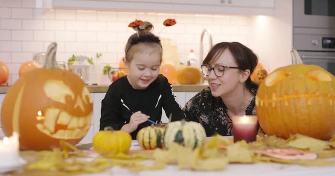 Woman in glasses sitting at kitchen table and looking at her daughter painting small pumpkin with gouache.