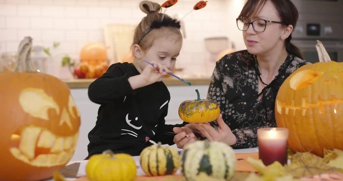 Smiling woman in glasses holding small painted pumpkin and showing it to her cute little daughter in Halloween costume.