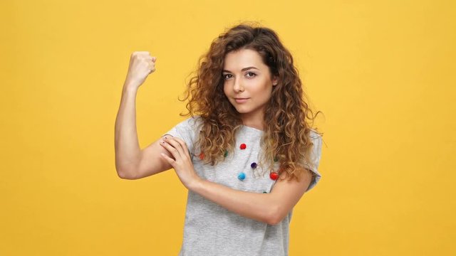 Smiling curly woman in t-shirt showing her bicep and looking at the camera over yellow background