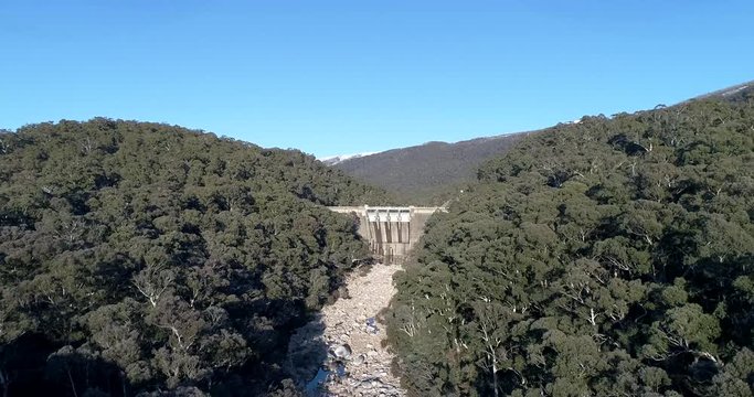 Snowy river flying from riverbed to Hydro dam between evergreen gumtrees under blue sky.
