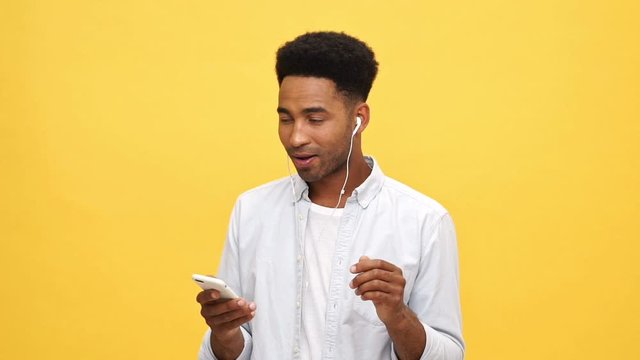 Happy african man in shirt listening music by headphones and dancing while using his smartphone over yellow background
