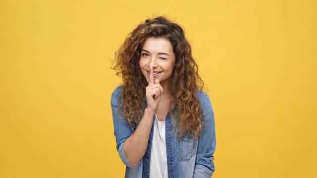 Smiling sly curly woman in denim shirt showing silence sign and winks at camera over yellow background