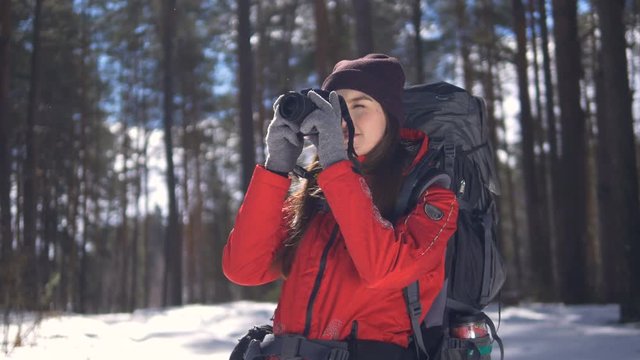 Smiling happy young woman hiking in winter forest taking pictures using photocamera.