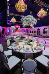 Large white bouquets of roses stand on dinner tables decorated with golden glasses and violet cloth