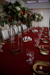 Tall bouquets of red flowers and greenery stand on dinner table covered with red cloth
