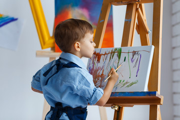 Cute, serious and focused, three years old boy in blue shirt and jeans apron painting on canvas standing on the easel. Concept of early childhood education, talent, happy family or parenting