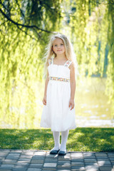 Cute little blonde girl is backlit against the background of water and grass tree