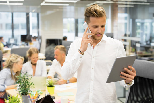 Man in open office talking on the phone and looking at tablet