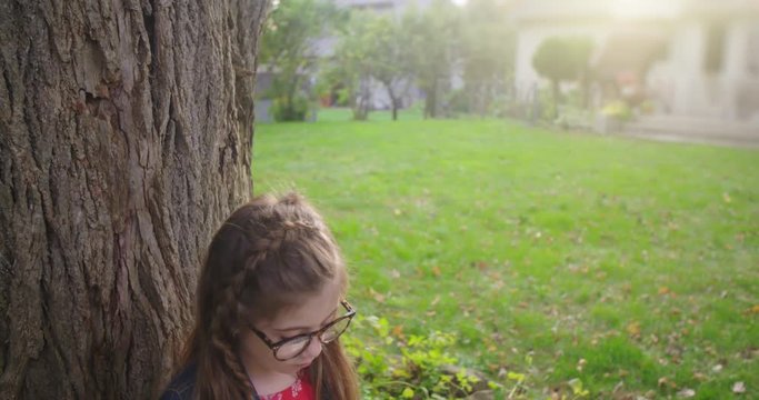 Young Girl with Book Shushes at Camera to be Quiet. a lowering shot towards a little girl with glasses reading a book under a tree when she turns to camera and shushes to be quiet
