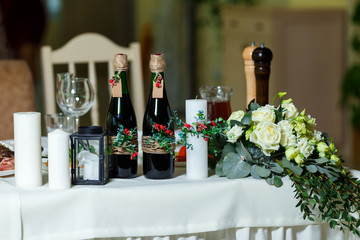 The wide white candle and two green bottles with red wine decorated with flowers, greenery and twine. Wedding table setting and decor