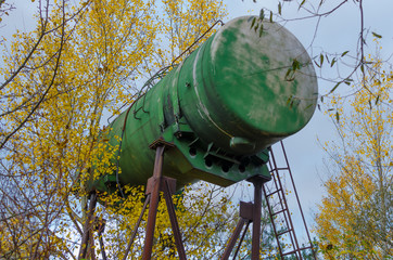 Huge green aluminium barrel on rusty metal support used as water tower near Grabovka village, Gomel region, Belarus, with autumn trees with yellow falling leaves and foggy blue sky.