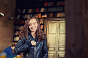 Film effect. Portrait of attractive young brunette female student leaving modern cafeteria or anti cafe dressing up leather jacket and smiling looking forward. Indoor shot of cute lady with long hair.