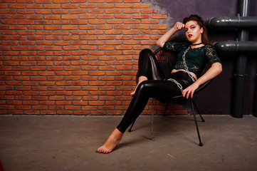 Black girl in green shirt and leather pants with bright make-up sitting at chair on studio background brick wall. Halloween theme.