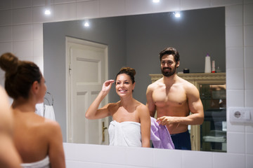 Young cute girl plucking eyebrows depilating with tweezers in front of a strong muscular shirtless man whole holding towel in the bathroom.