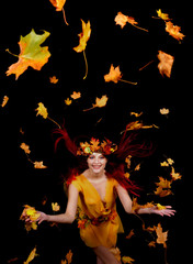 A beautiful young woman dresses up as an Autumn fairy   and wears a wreath on her head made out of Autumn leaves.  She poses for the camera while Autumn leaves fly around her.