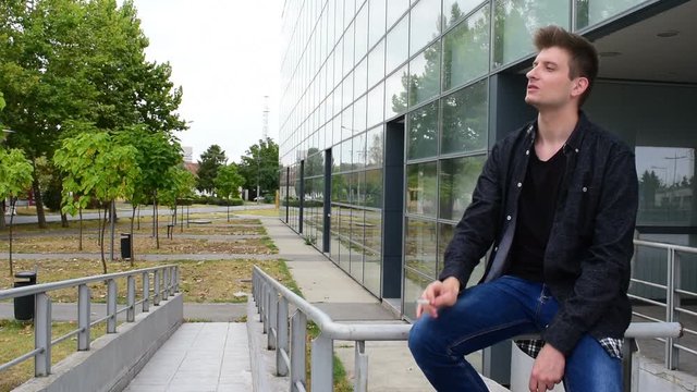 casual young man sitting on a guardrails against the glass building, smokes a cigarette