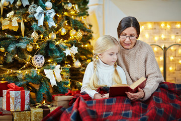 Aged woman reading book of fairy-tales to cute girl by decorated Christmas tree