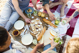 Various food on picnic table and several friends around it eating sandwiches and fruit and drinking homemade lemonade