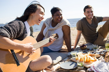 Joyful young man playing guitar in circle of friends during picnic by seaside