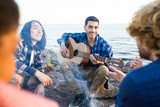 Happy guy with guitar and his friends spending evening around campfire by seaside