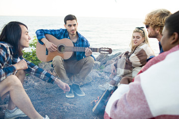 Friends sitting in circle aroung campfire by waterside in the evening and singing by guitar