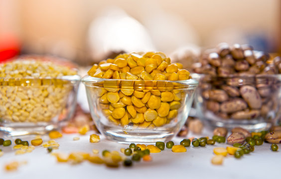 Assorted lentils and beans in glass bowls, featuring vibrant red gram (toor dal), split moong dal, and speckled rajma beans. Scattered whole legumes such as green gram and black gram add to the visual