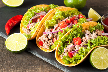 Photo of Mexican tacos with ground meat, beef, beans, onions and salsa on wooden background. Ketchup sauce and lime. A glass o beer in the background.