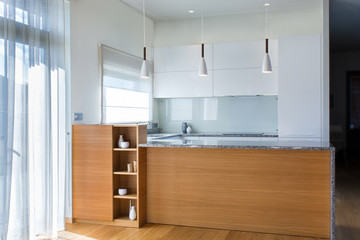 Modern kitchen furniture design in light interior with wood accents. Kitchen peninsula in the room. Stone surface. Gloss facades. Finished project. White pendant light. Sink, window, blinds. Glass.