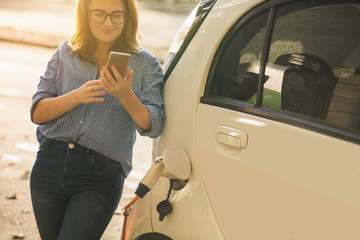 Young woman is standing near the electric car and looks at the smartphone. The rental car is charging at the charging station for electric vehicles. Car sharing.