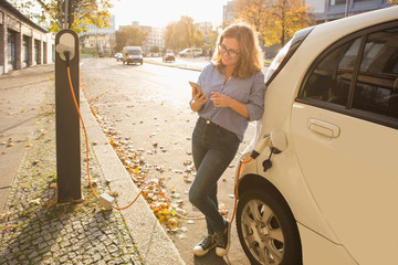 Young woman is standing near the electric car and holding smartphone. The rental car is charging at...