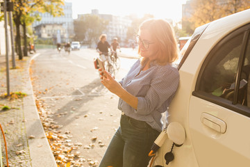Young woman is standing near the electric car and holding smartphone. The rental car is charging at...