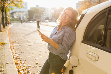 Young woman is standing near the electric car and holding smartphone. The rental car is charging at the charging station for electric vehicles. Car sharing.