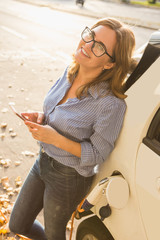 Young woman is standing near the electric car and holding smartphone. The rental car is charging at...